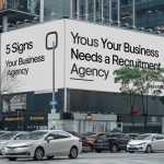 A photo of a busy city street with a large billboard that has the text 5 Signs Your Business Needs a Recruitment Agency. The billboard is located on a building and is the main object in the photo. The background contains cars, trees, and other buildings.