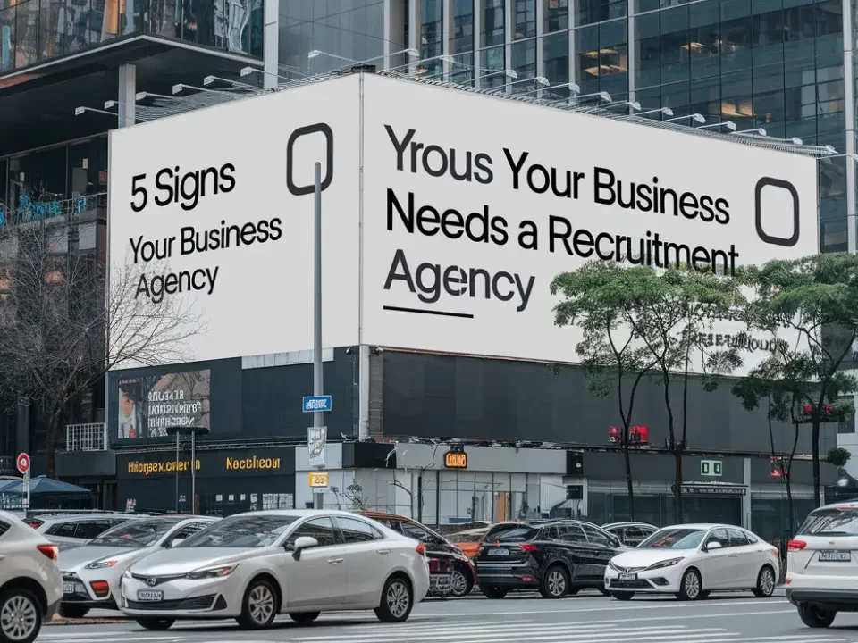 A photo of a busy city street with a large billboard that has the text 5 Signs Your Business Needs a Recruitment Agency. The billboard is located on a building and is the main object in the photo. The background contains cars, trees, and other buildings.